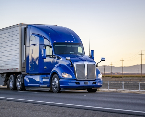 Industrial comfort big rig powerful blue semi truck tractor with high cab transporting frozen commercial cargo in refrigerator semi trailer running on the highway road at sunset in California