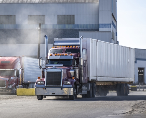 Pro commercial classic burgundy big rig semi truck tractor with chrome parts and container on semi trailer running on the road in industrial area raising a cloud of dust