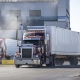 Pro commercial classic burgundy big rig semi truck tractor with chrome parts and container on semi trailer running on the road in industrial area raising a cloud of dust