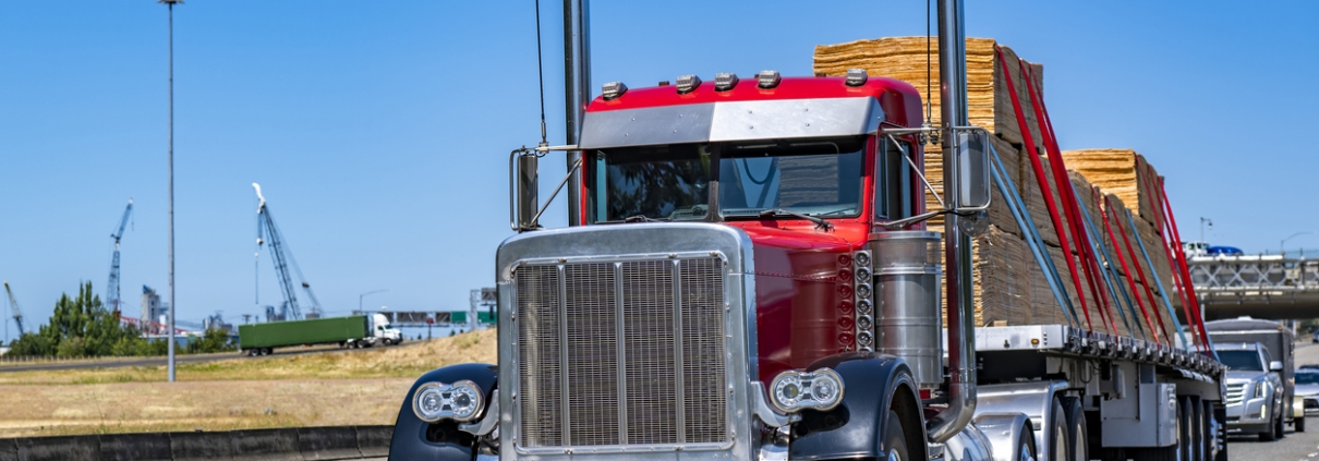Industrial carrier red day cab big rig classic old style semi truck tractor with chrome parts transporting fastened plywood on flat bed semi trailer running on the wide highway road at sunny day