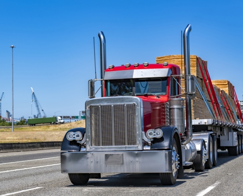 Industrial carrier red day cab big rig classic old style semi truck tractor with chrome parts transporting fastened plywood on flat bed semi trailer running on the wide highway road at sunny day