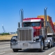 Industrial carrier red day cab big rig classic old style semi truck tractor with chrome parts transporting fastened plywood on flat bed semi trailer running on the wide highway road at sunny day