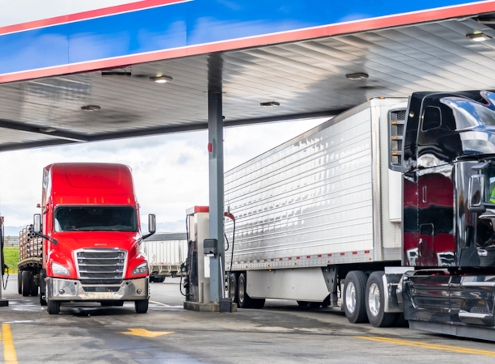 Industrial standard big rigs semi trucks with semi trailers standing on the fuel station parking lot filling the semi trucks tanks with diesel fuel to have ability continue commercial flight