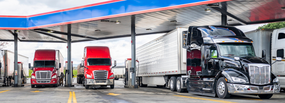 Industrial standard big rigs semi trucks with semi trailers standing on the fuel station parking lot filling the semi trucks tanks with diesel fuel to have ability continue commercial flight