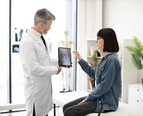 Confident mature man in lab coat holding digital tablet while young lady in cervical collar sitting in exam room. General practitioner showing test results to female with car accident injury.